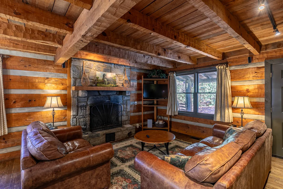 A warm and inviting living room featuring a substantial stone fireplace as the focal point. Plush leather sofas are arranged for cozy conversation or lounging, with wooden beams overhead lending rustic charm. The mantel is adorned with decorative pieces, and a TV sits nearby, hinting at modern comforts.