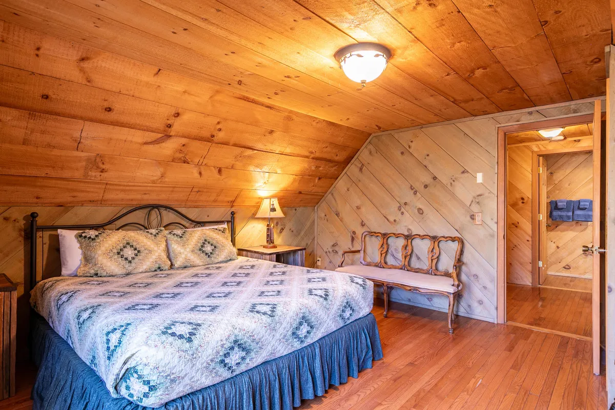 An inviting bedroom with a cozy bed draped in a patterned quilt, a wooden ceiling, a nightstand with a lamp, and a bench set against the wall. The soft lighting and decor give the room a charming, antique feel—ideal for family or friends seeking privacy and comfort upstairs.