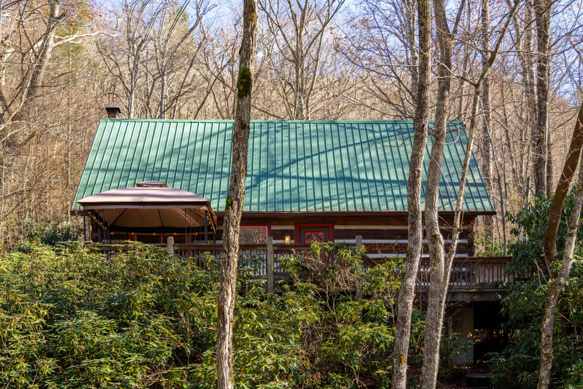 The image provides a wide view of the log cabin, emphasizing the green metal roof, spacious wooden deck, and the surrounding trees. The deck area suggests an ideal space for gathering with friends or simply taking in the beauty of the mountains.