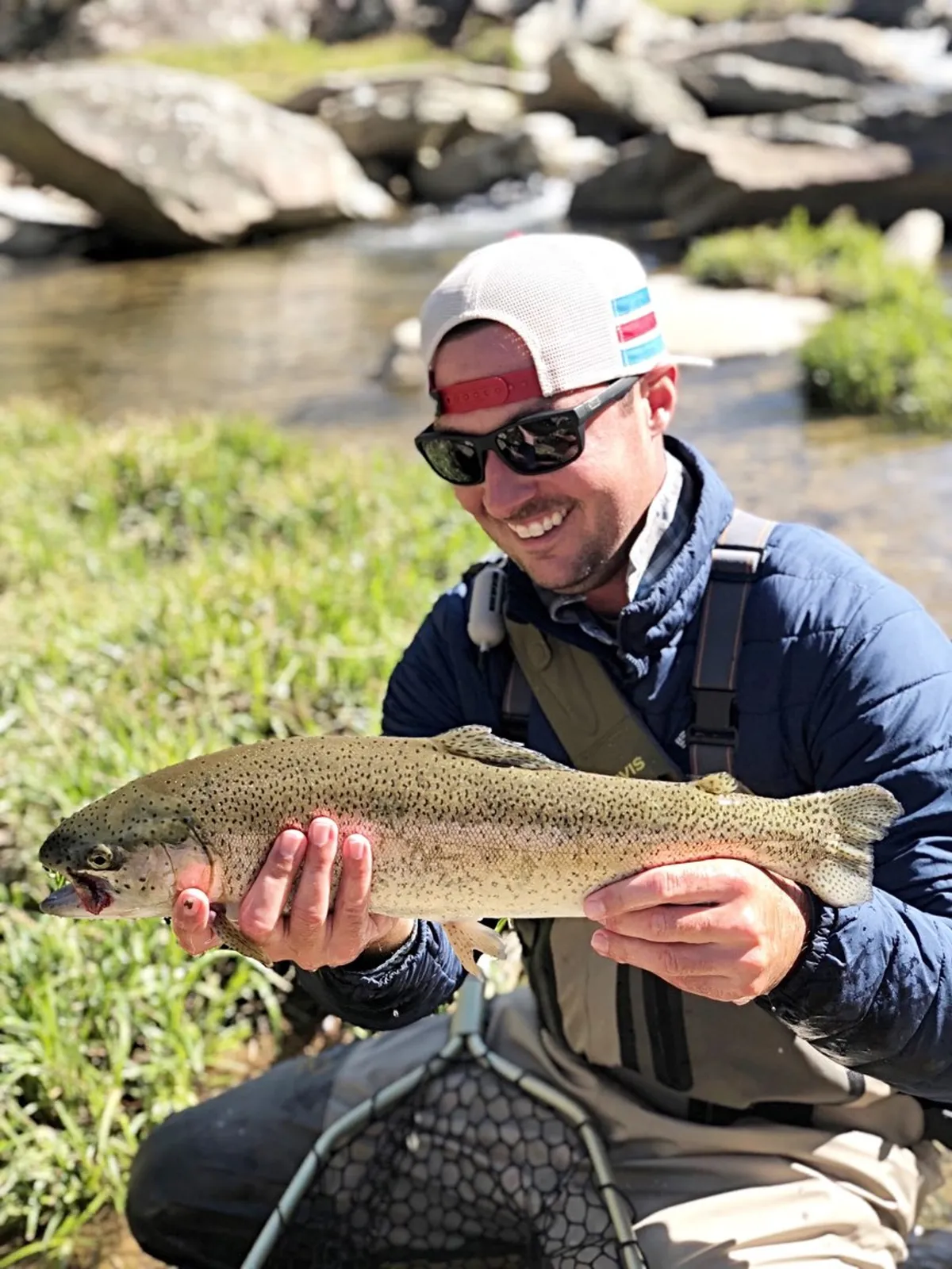 A man proudly holds up a large fish beside a crystal-clear stream and rocky banks. The joyful scene highlights the thrill of fishing and the beauty of the outdoors, with verdant greenery as the backdrop.
