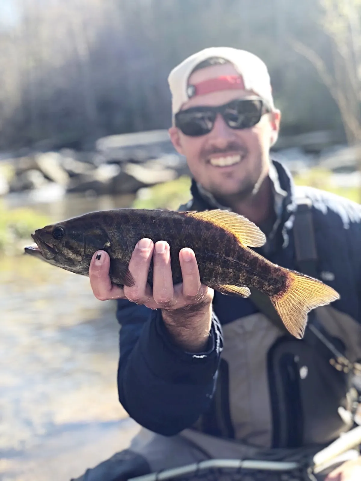 A person proudly holds a freshly caught fish beside a sparkling waterway, sunlight glinting off the river's surface. The surrounding setting is natural and vibrant, suggesting a successful and memorable day of fishing.