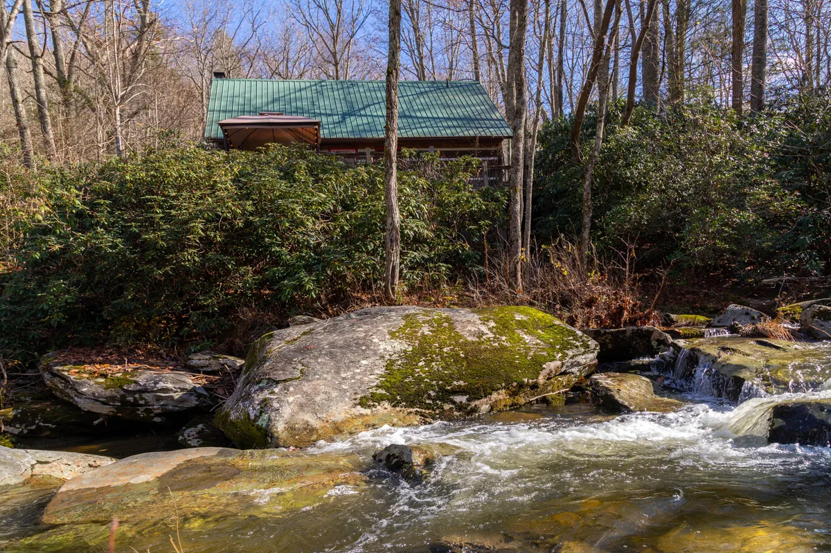 The hero image captures a charming log cabin nestled alongside the flowing Watauga River, surrounded by lush trees and natural rock formations. The tranquil stream sparkles in the foreground, with inviting outdoor spaces perfect for sipping morning coffee or relaxing after a day in the mountains. Sunlight filters through the canopy, enhancing the peaceful, secluded atmosphere of this mountain retreat.