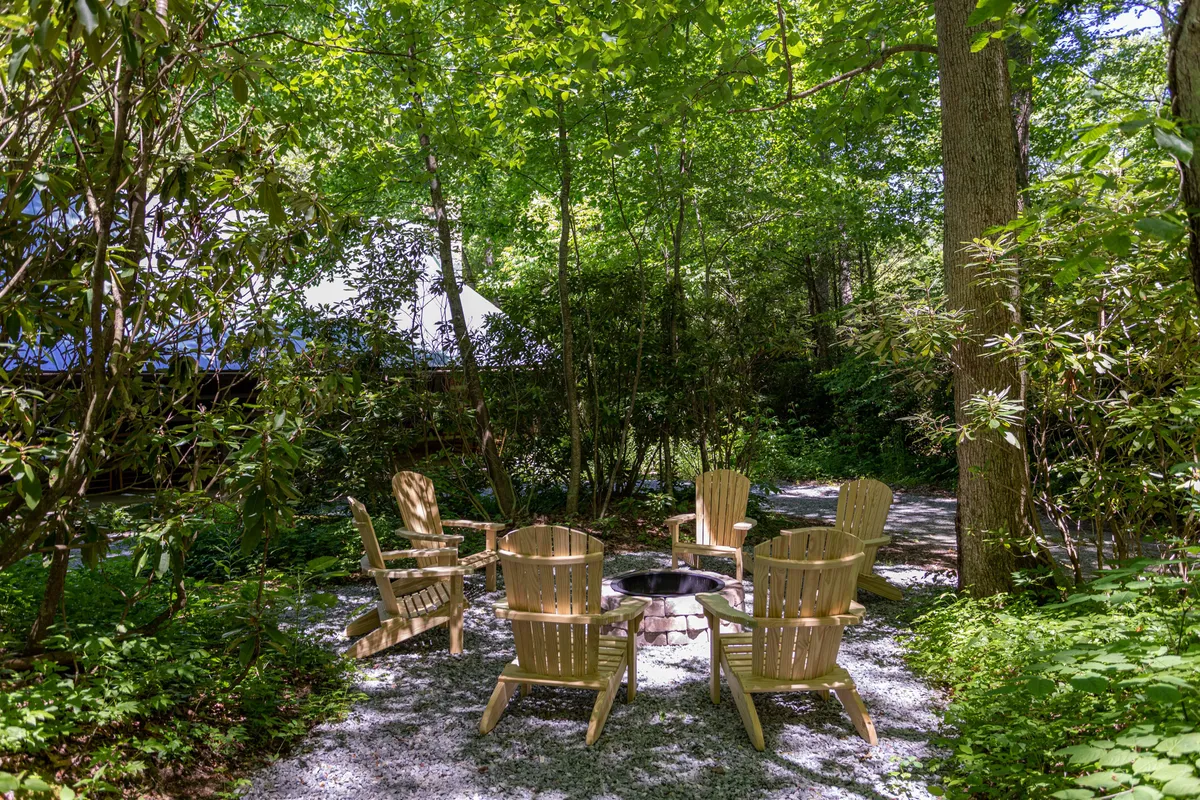 An exterior setup of a cozy fire pit circled by Adirondack chairs and surrounded by lush greenery. This inviting area is ideal for couples to snuggle, stargaze, or share stories under the night sky.