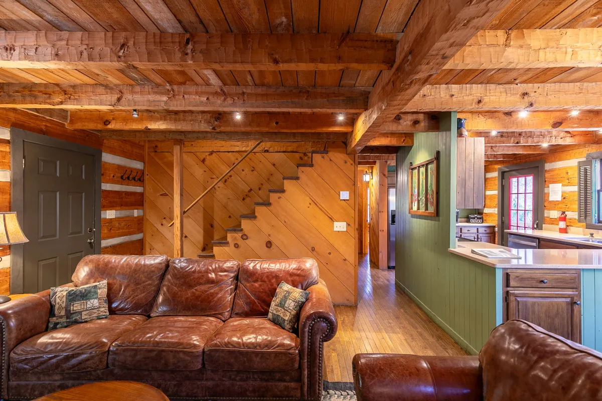 A broad view of the open-plan main living space with a leather sofa, visible wooden staircase, kitchen area in the background, and wall art. The scene feels lively and social, emphasizing spaces for relaxation and connection.