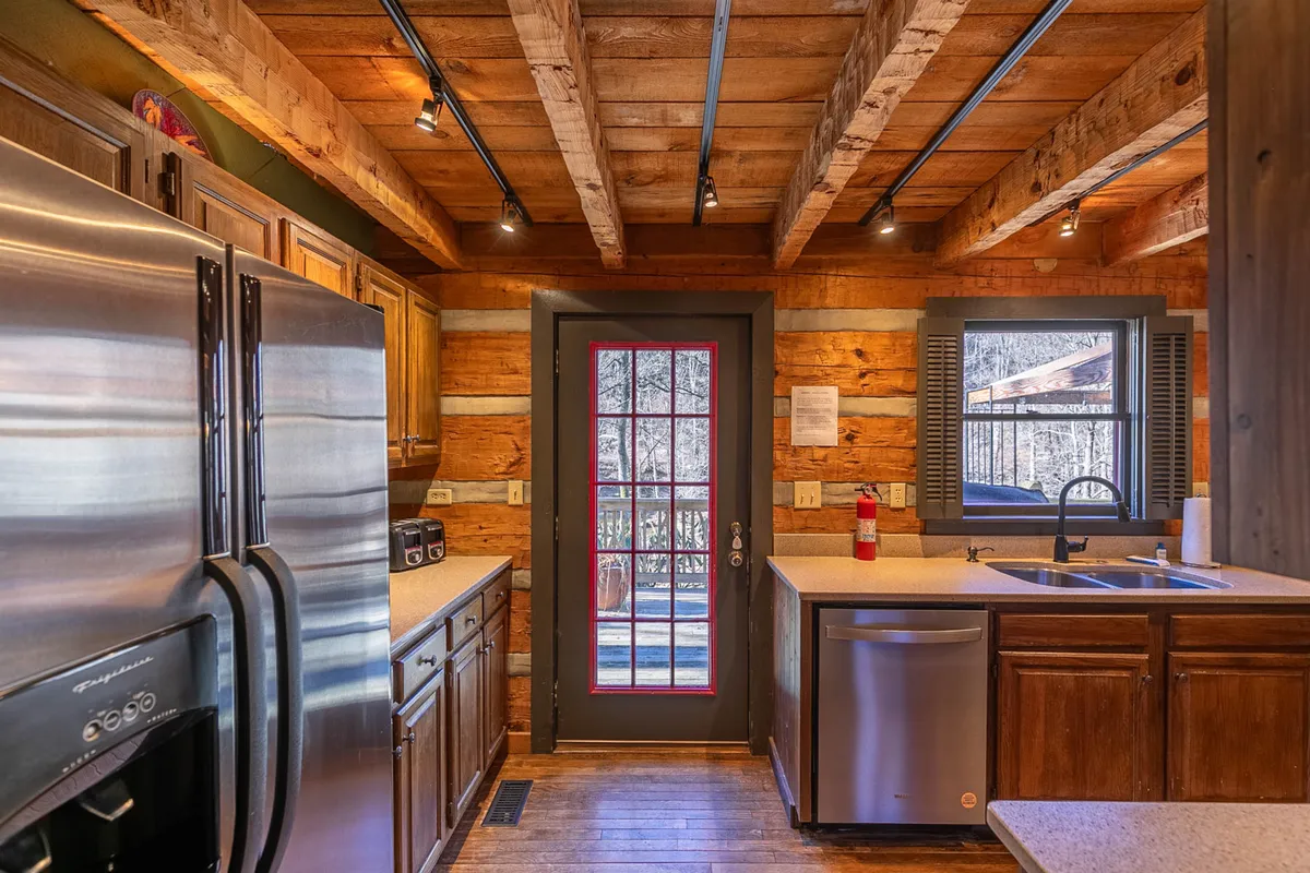 A spacious, sun-filled kitchen with stainless steel appliances, wooden cabinetry, exposed beam ceilings, and a large window. The scene evokes a welcoming atmosphere, ideal for family gatherings and culinary activities. The kitchen island and functional layout make it the heart of the home, inviting everyone to participate in meal prep and conversation.