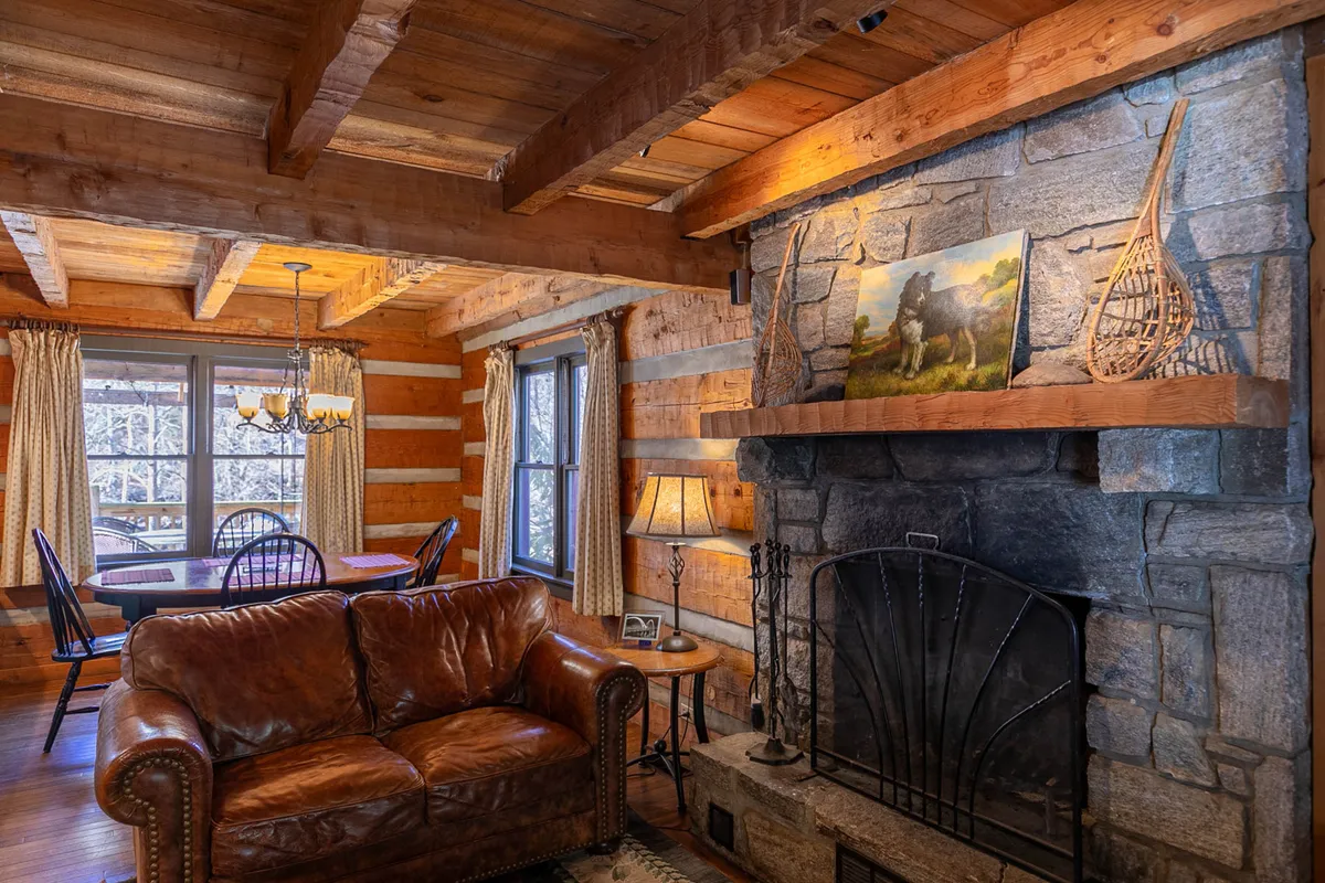 A view of the living and dining area, showcasing a crackling stone fireplace, leather sofa, sturdy wooden beams, and original painted artwork. The dining table is visible, emphasizing the open layout and welcoming atmosphere for group gatherings.