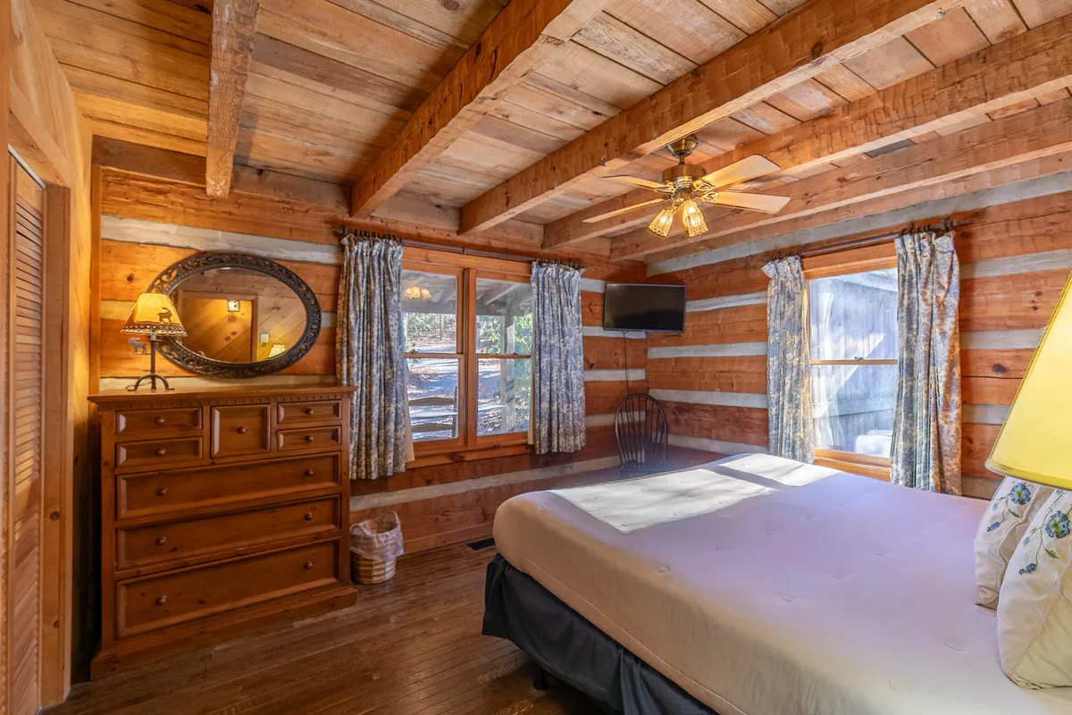 A spacious bedroom featuring wooden ceiling beams, a dresser with a mirror, large windows allowing in natural light, a comfortable bed with decorative pillows, and a ceiling fan. The room feels both inviting and luxurious, with views of nature visible through the windows.