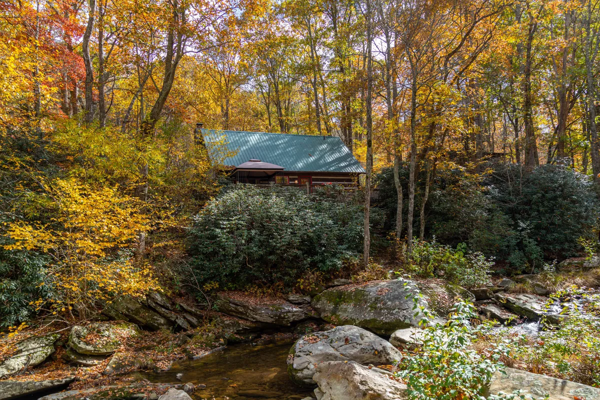 A tranquil scene featuring a cozy log cabin nestled among tall trees and brilliant autumn foliage, with a gently flowing stream in the foreground. The cabin’s exterior radiates seclusion and warmth, making it an inviting haven for couples seeking privacy and natural beauty.